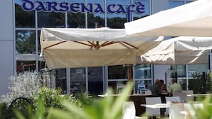Un área de café al aire libre con mesas y sillas blancas, un toldo grande y un edificio con un letrero que dice "DARSENACAFE".