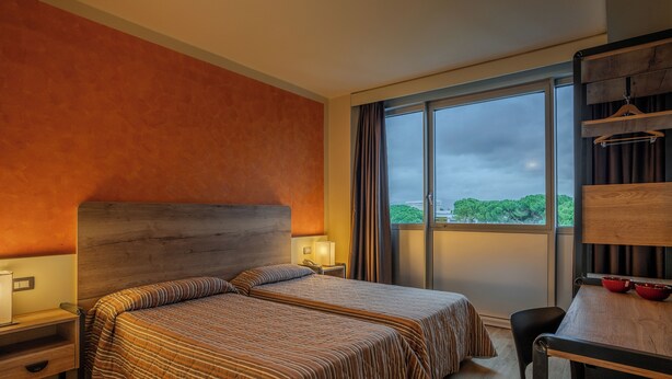 A hotel room with two beds, a desk, and a large window with a view of greenery. - Hotel Galilei (Pisa)