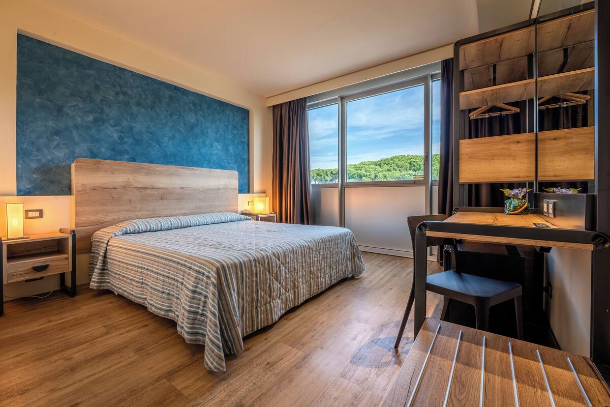 Habitación de hotel con cama, mesitas de noche, escritorio y una silla. Amplia ventana con vistas a zonas verdes.