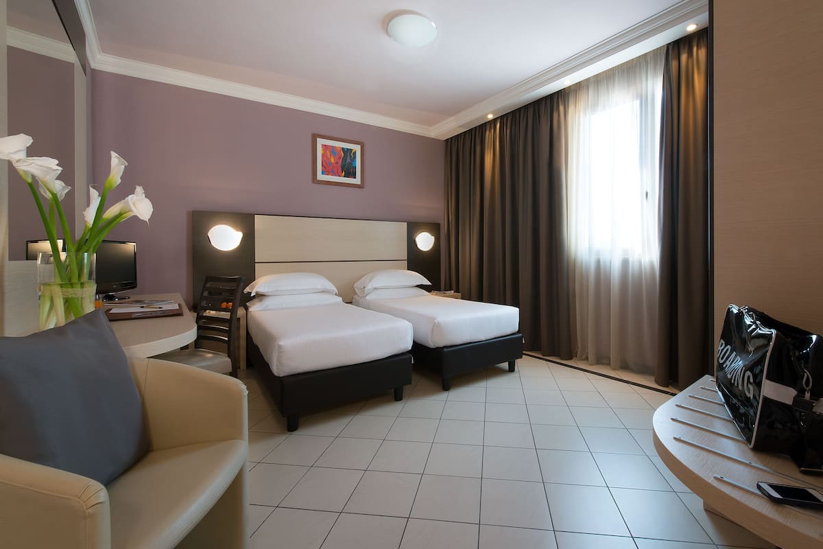 A hotel room with two beds, a desk, a chair, a TV, and a vase with flowers.