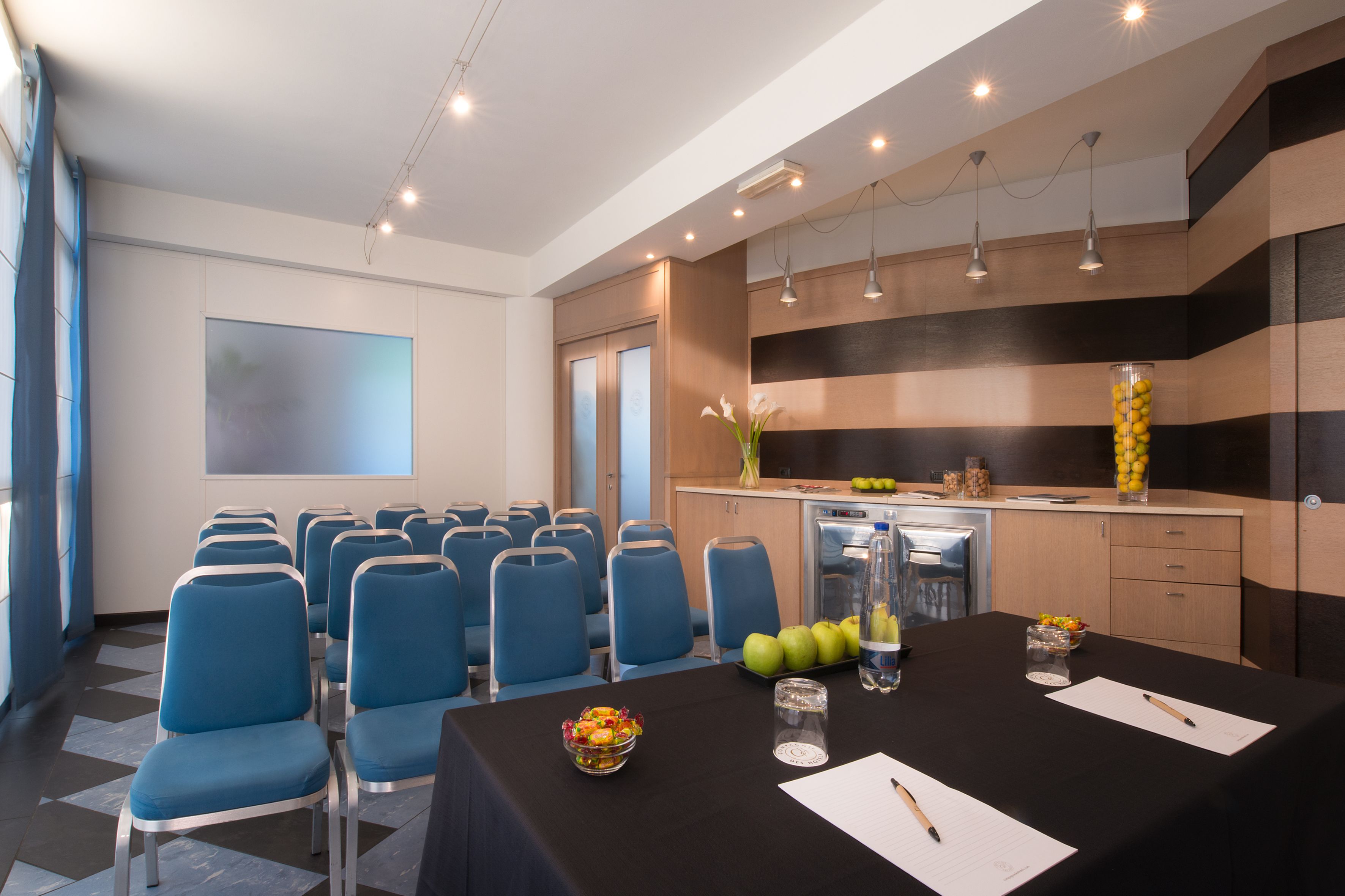 A conference room with blue chairs, a black table with a bowl of fruit, a water bottle, and a projector screen.