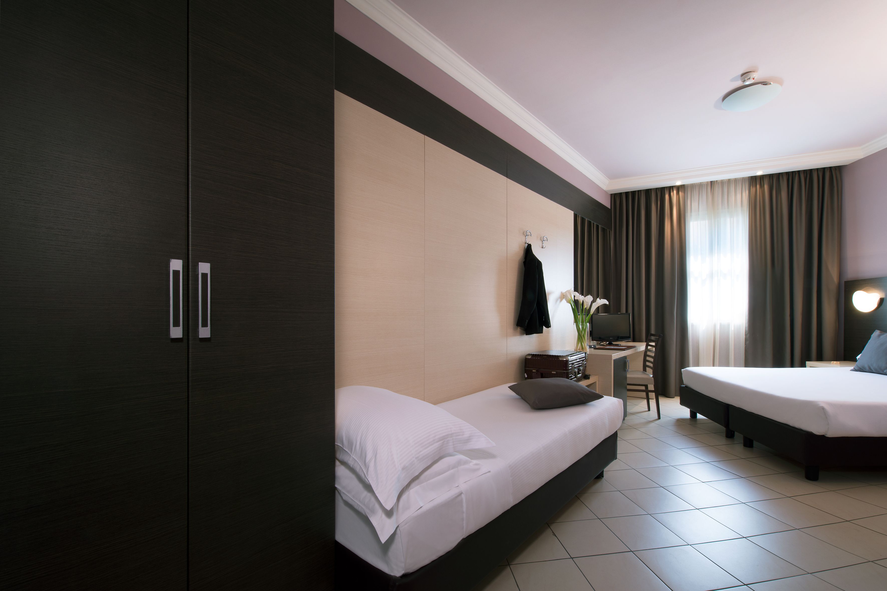 A hotel room with a large wardrobe, two beds, a desk with a chair, and a window with curtains.