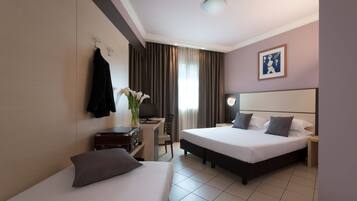 A hotel room with two beds, a desk, a chair, a desk lamp, a computer, a vase with flowers, and a framed picture on the wall.
