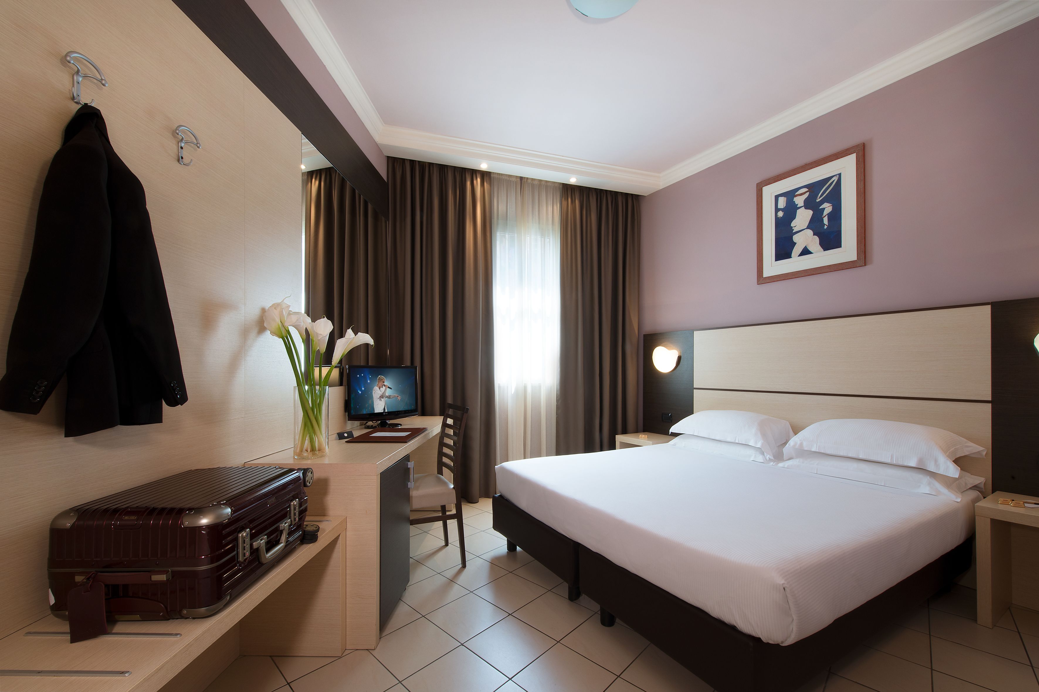 Une chambre d’hôtel avec un lit, un bureau avec un ordinateur, une chaise, une valise et un porte-manteau.