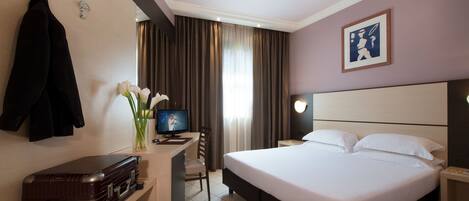 Une chambre d’hôtel avec un lit, un bureau avec un ordinateur, une chaise, une valise et un porte-manteau.