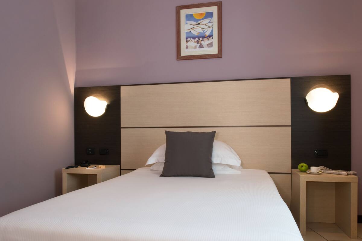 A neatly made bed with a white comforter and a dark pillow, flanked by two bedside tables with lamps, one of which has a green apple on it.