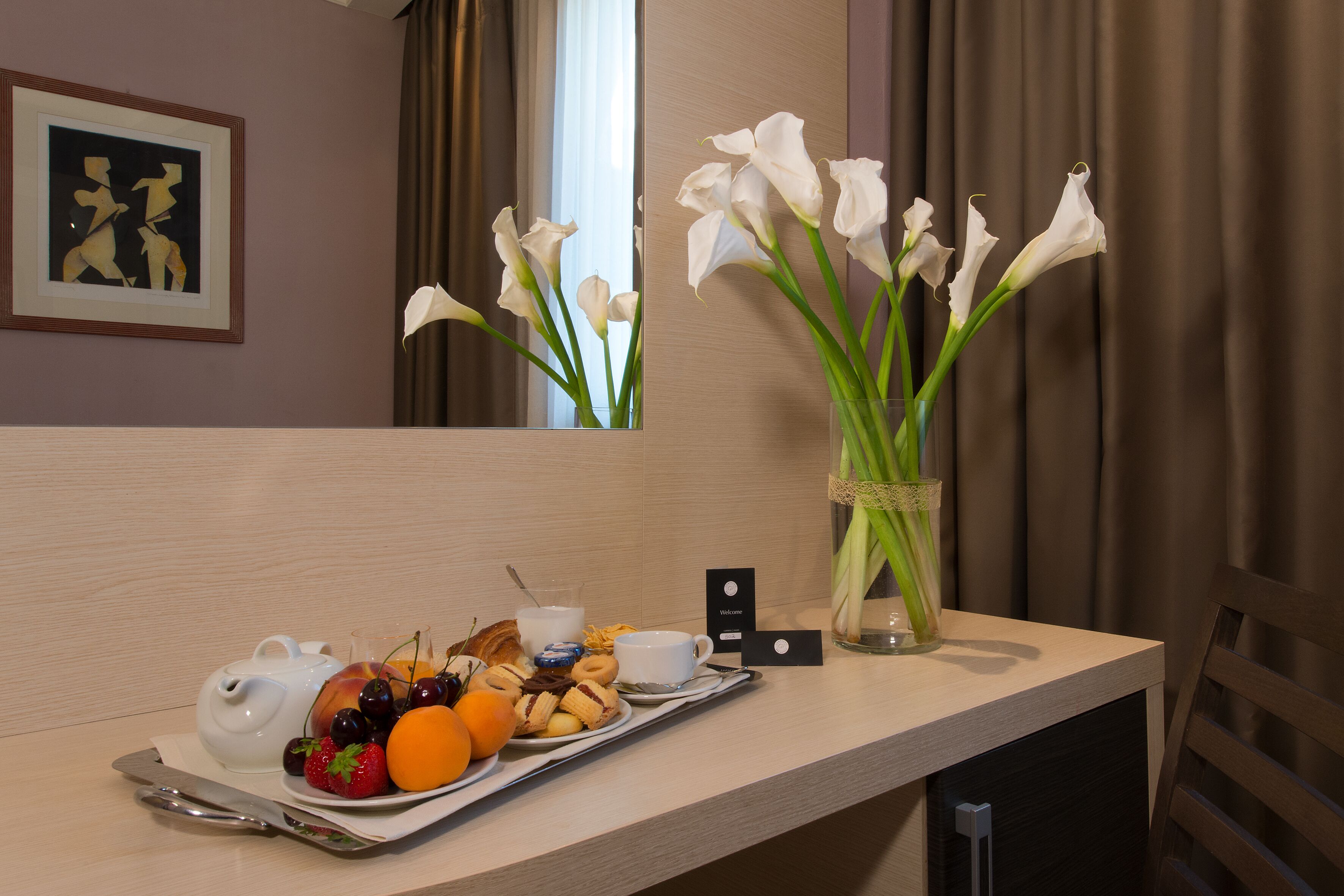 Une chambre d’hôtel avec un plateau de petit-déjeuner, un vase de fleurs blanches et une œuvre d’art encadrée accrochée au mur.