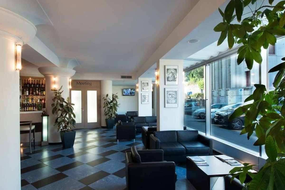 A hotel lobby with checkered flooring, seating areas, a bar, and large windows offering a view of parked cars.