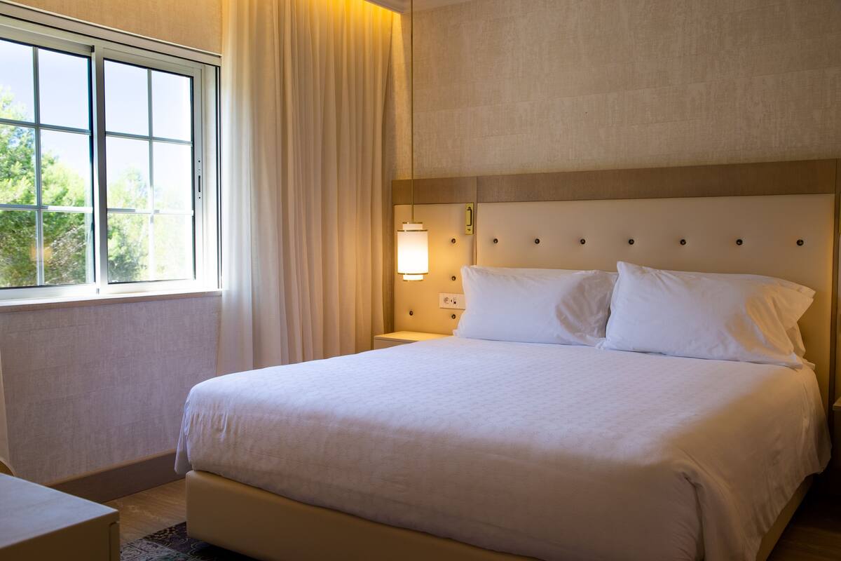 A hotel room with a large bed, a bedside table, a nightstand, a window with curtains, and a wall-mounted lamp.