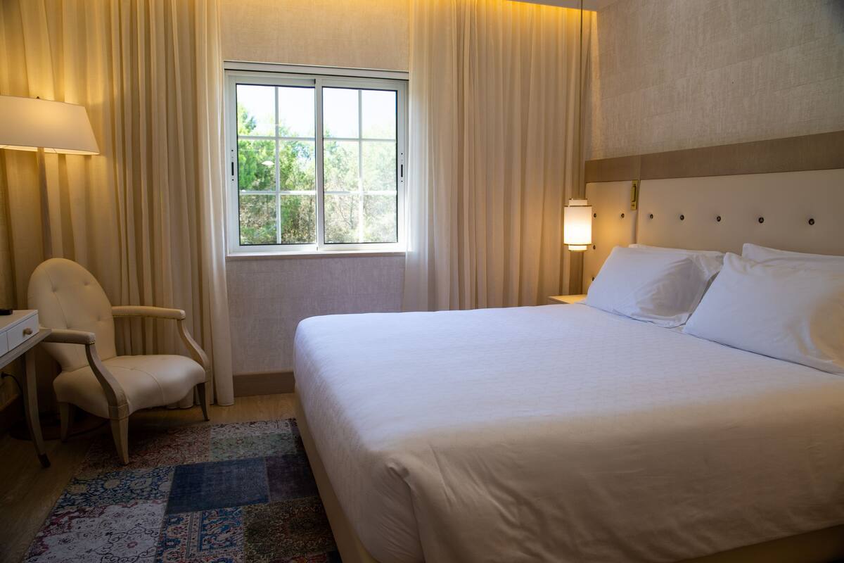 A hotel room with a large bed, a desk, a chair, a window with curtains, and a nightstand with a lamp.