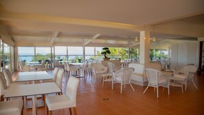 A spacious dining area with white tables and chairs, large windows offering a view of the sea, and a central plant display. - Vila Alba Resort (Lagoa)