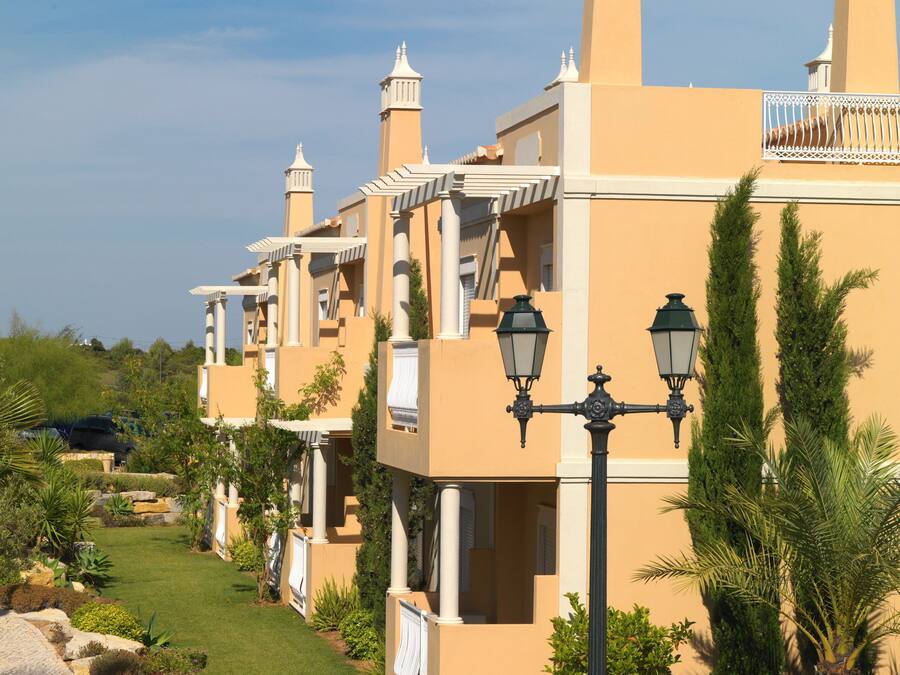 A row of beige buildings with white balconies, a paved pathway, and landscaped gardens.