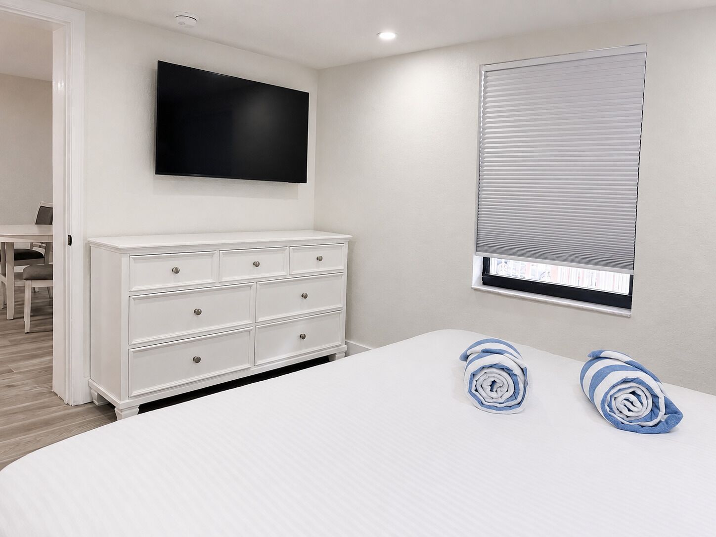 Ein Schlafzimmer mit einem Bett, einer weißen Kommode, einem flachbildfernseher an der Wand und einem Fenster mit Jalousien.
