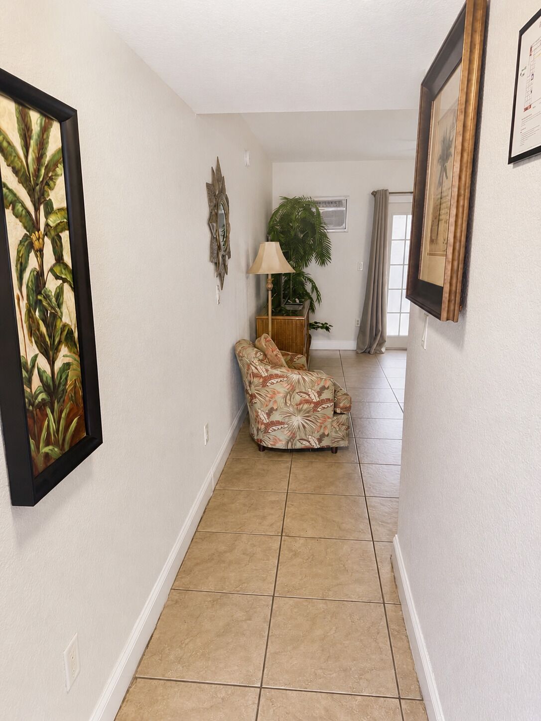 A hallway with a tiled floor, a chair with a floral pattern, a lamp, and two framed pictures on the walls.