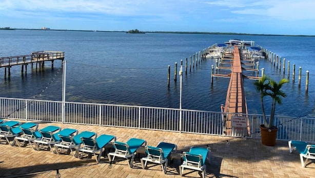 A marina with a wooden dock extending into calm waters, surrounded by a white railing and blue lounge chairs.