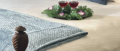 Una cama bien hecha con colcha azul y blanca, dos almohadas, una bandeja decorativa con dos copas de vino tinto y una manta azul.