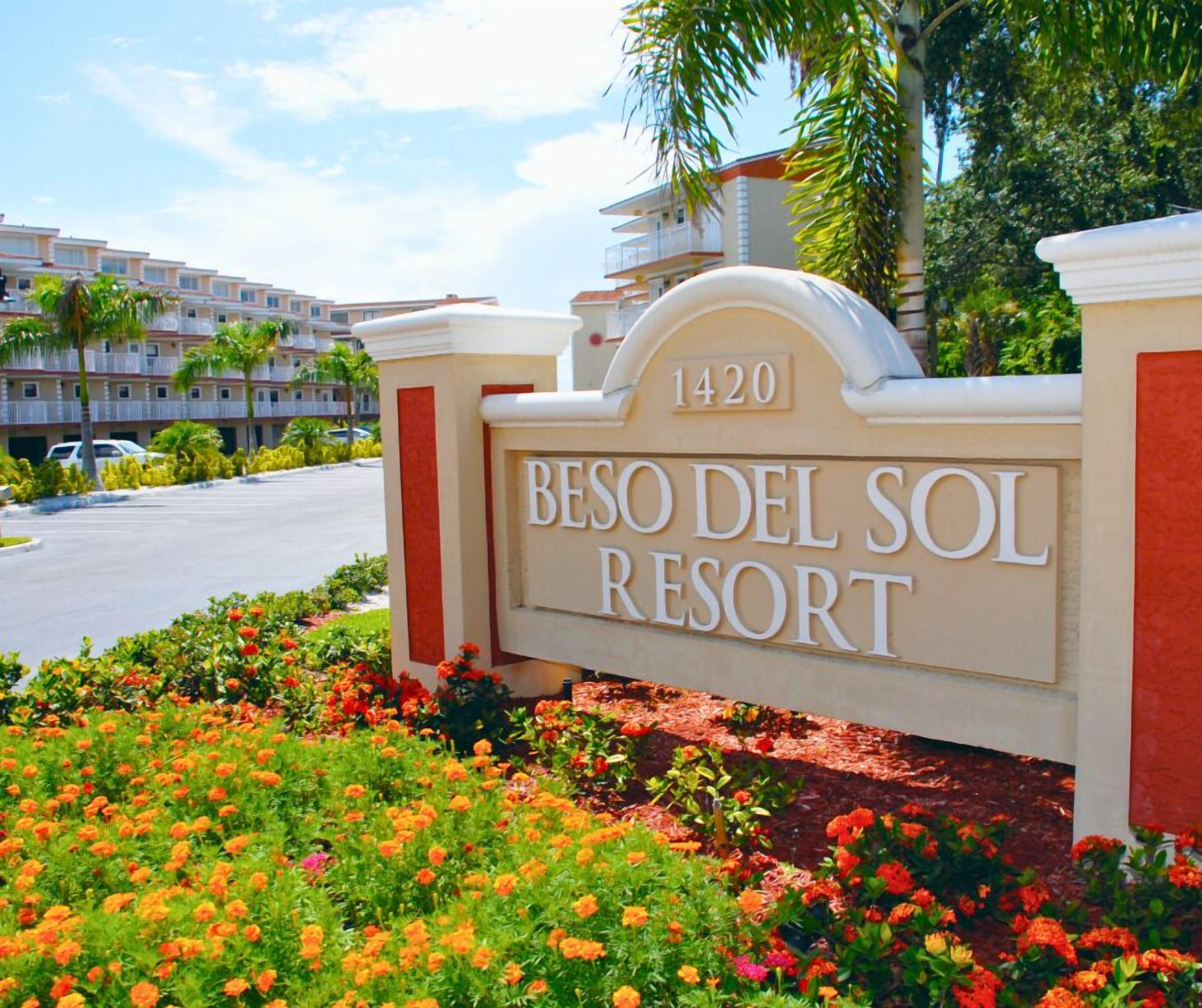 Letrero del RESORT BESO DEL SOL con el número 1420, rodeado de flores coloridas y palmeras.