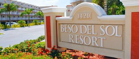 Letrero del RESORT BESO DEL SOL con el número 1420, rodeado de flores coloridas y palmeras.