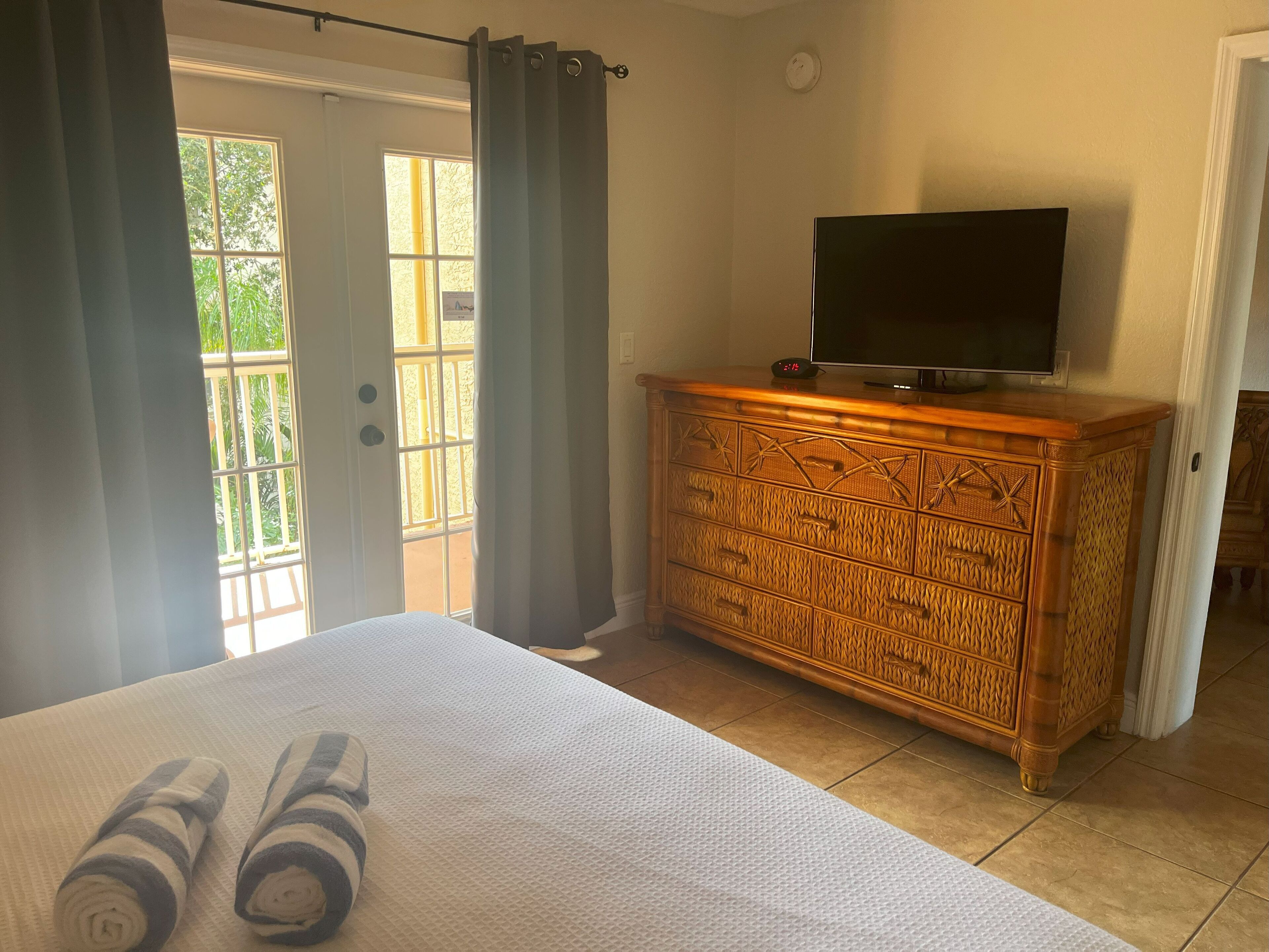 A hotel room with a bed, a wooden dresser with a television, and a view of the outdoors through a door.