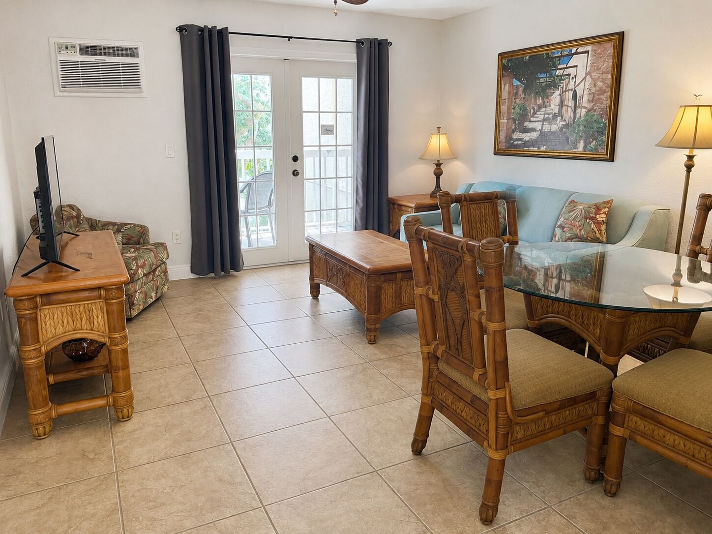 A living room with a glass-top dining table, wooden chairs, a sofa, a television, and a painting on the wall.