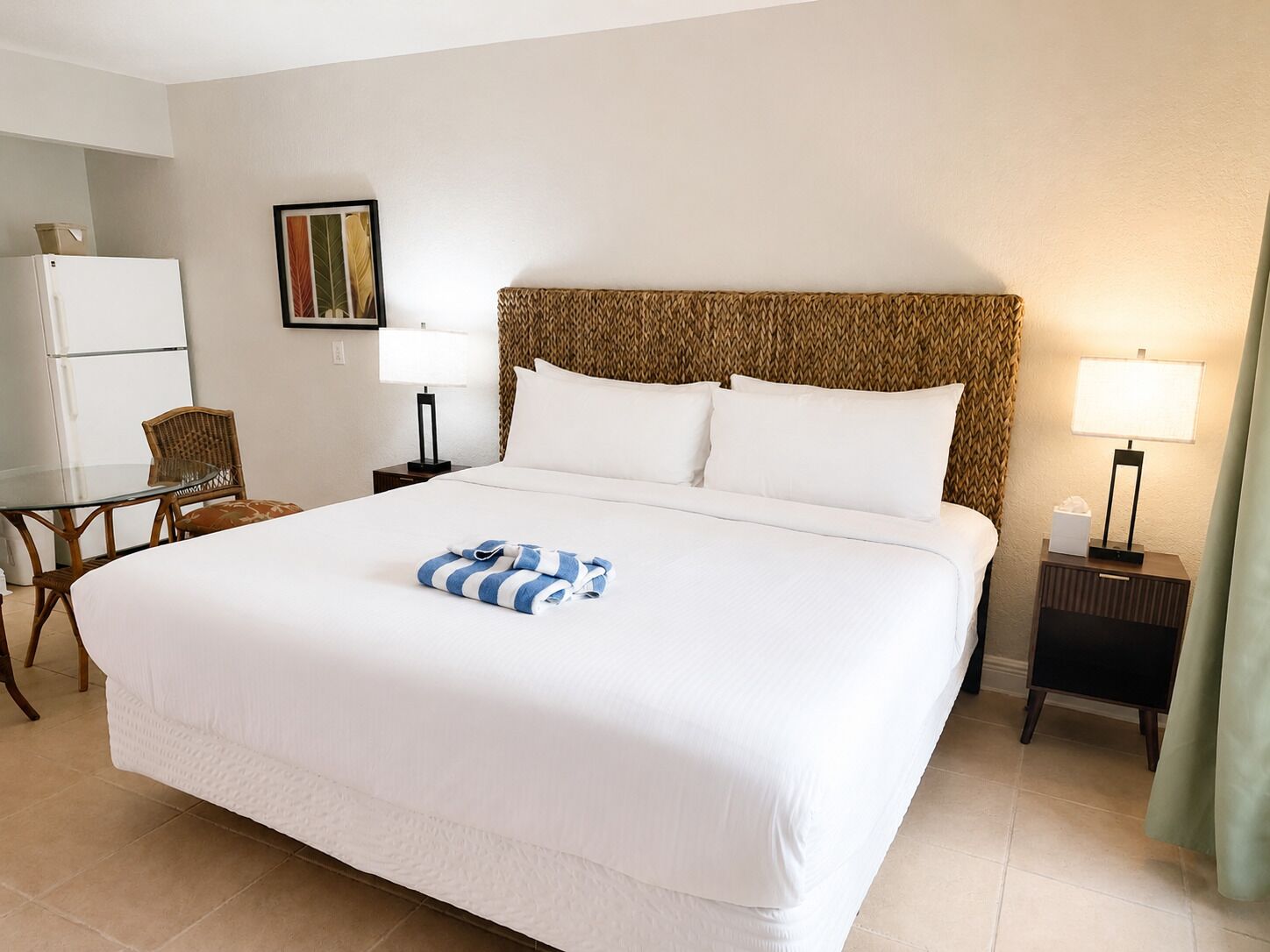 A hotel room with a large bed, two bedside tables, a chair, a refrigerator, and a painting on the wall.