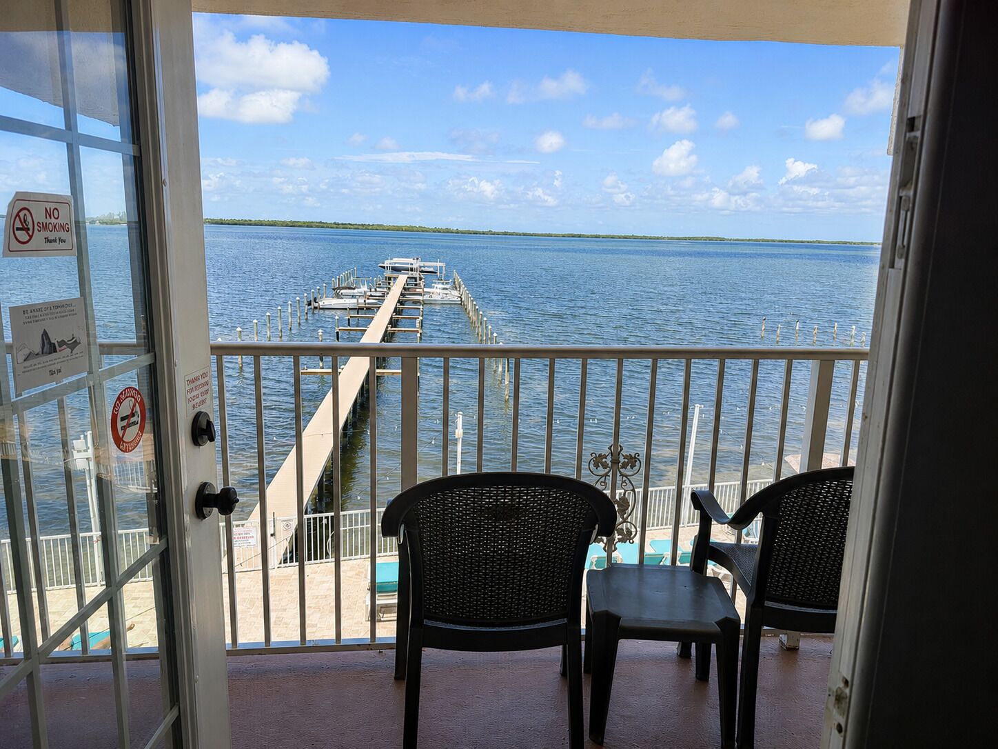 Un balcon avec deux chaises, une vue sur un quai s’avançant dans l’eau et un panneau « interdiction de fumer » sur la porte.