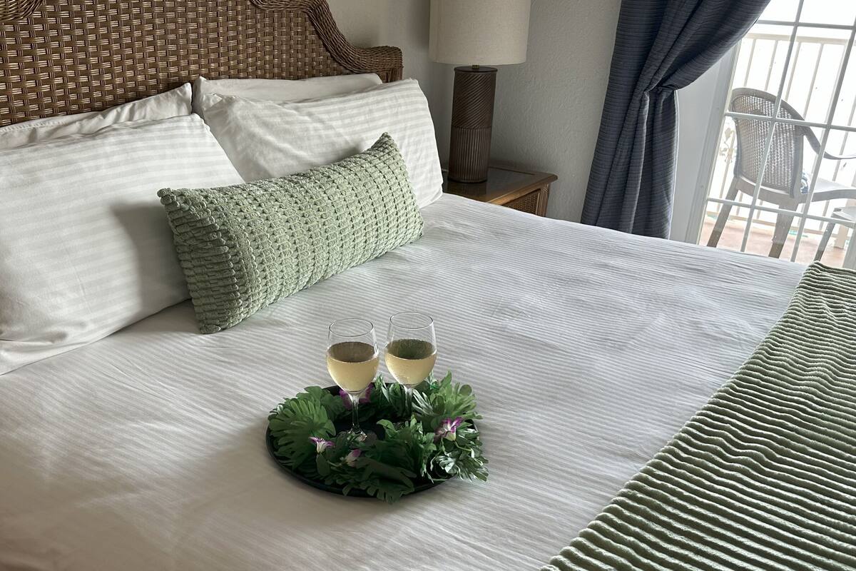 Un lit bien fait, avec du linge de lit blanc, deux verres de vin blanc posés sur un plateau décoratif et un coussin vert en tricot.