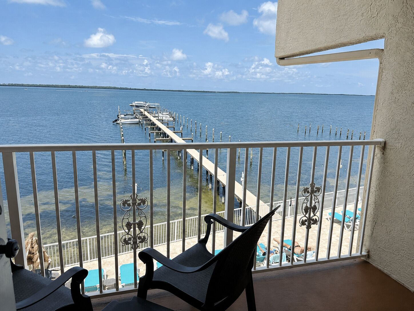Un balcon avec vue sur un quai et des bateaux, une piscine et des chaises longues.