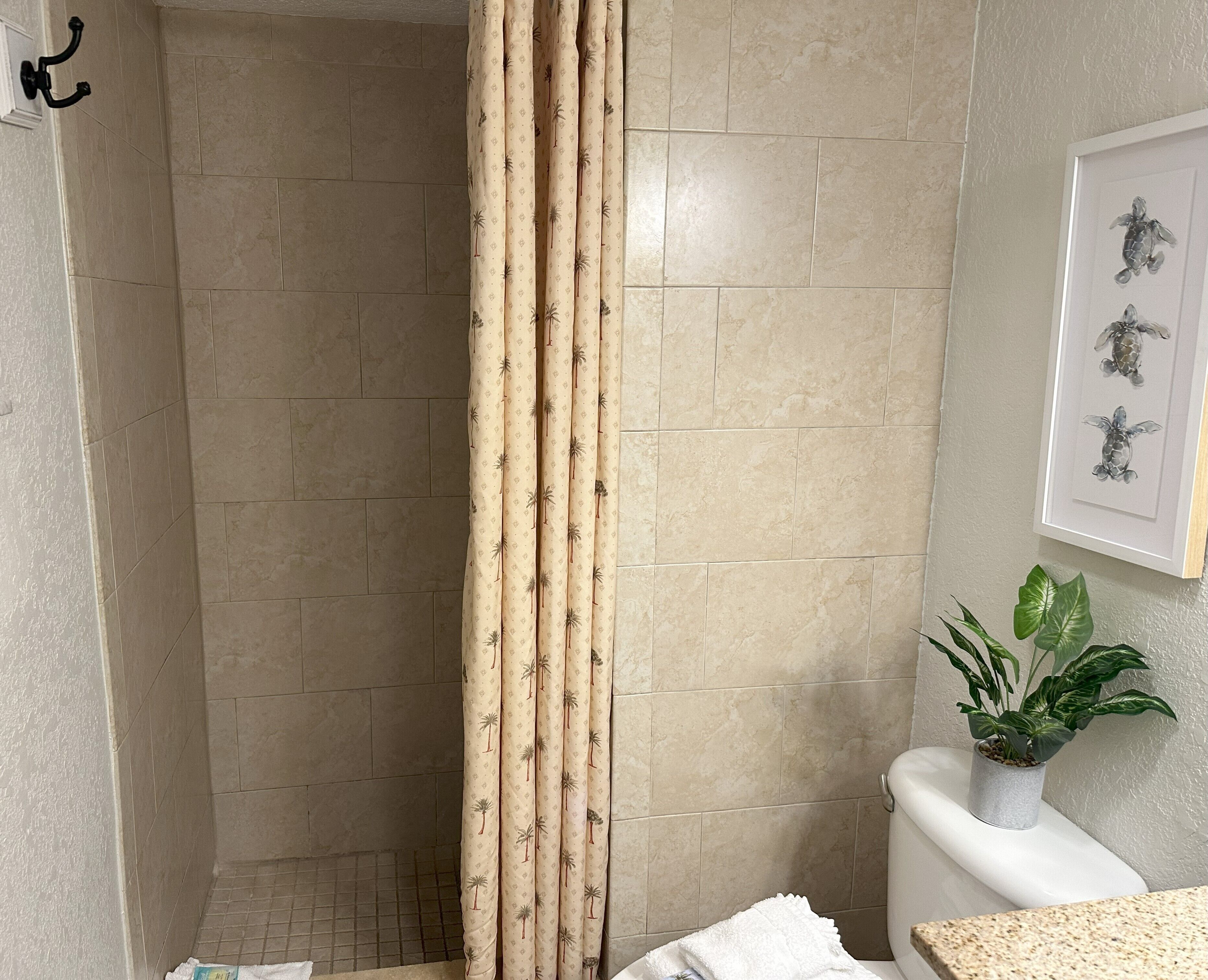 Un baño con azulejos color crema, una cortina de ducha con estampado de palmeras, un inodoro, un lavabo y un cuadro de una tortuga enmarcada en la pared.