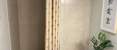 Un baño con azulejos color crema, una cortina de ducha con estampado de palmeras, un inodoro, un lavabo y un cuadro de una tortuga enmarcada en la pared.