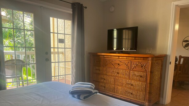Een slaapkamer met een bed, een houten dressoir, een televisie op het dressoir, en uitzicht op groen door een raam.
