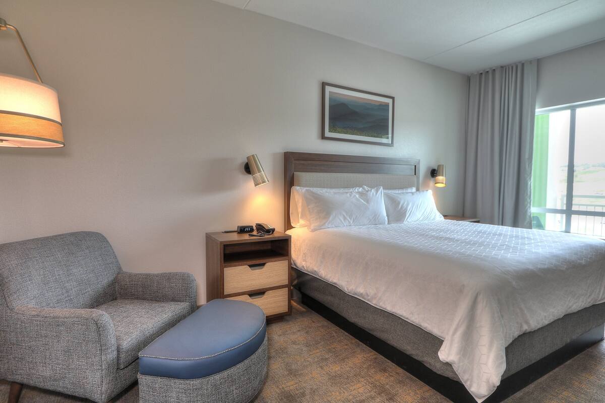 A hotel room with a bed, a chair, a nightstand, and a wall-mounted lamp.