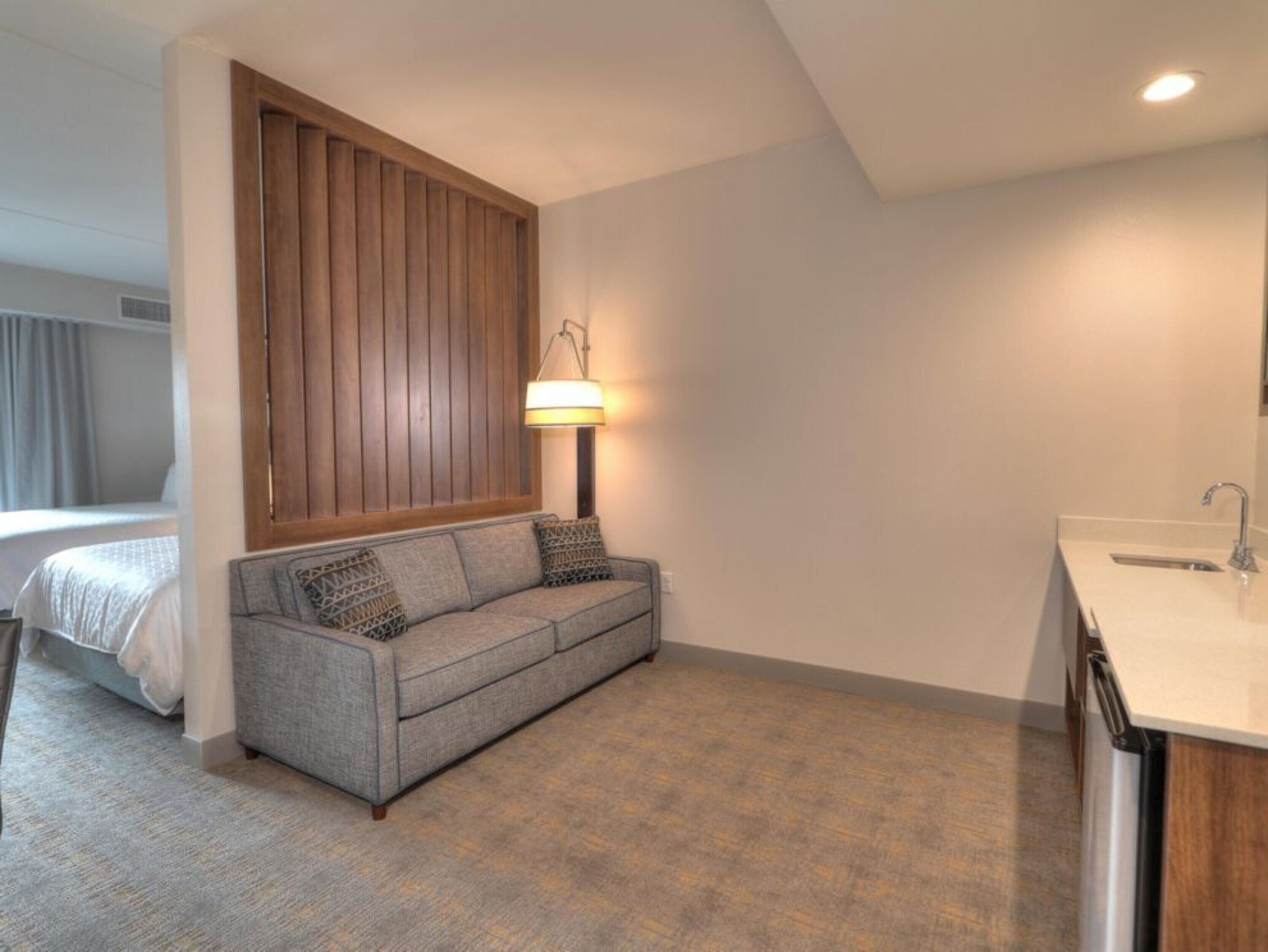 A modern hotel room with a grey sofa, a wooden panel wall, a bedside table, and a built-in sink.