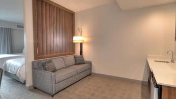 A modern hotel room with a grey sofa, a wooden panel wall, a bedside table, and a built-in sink.