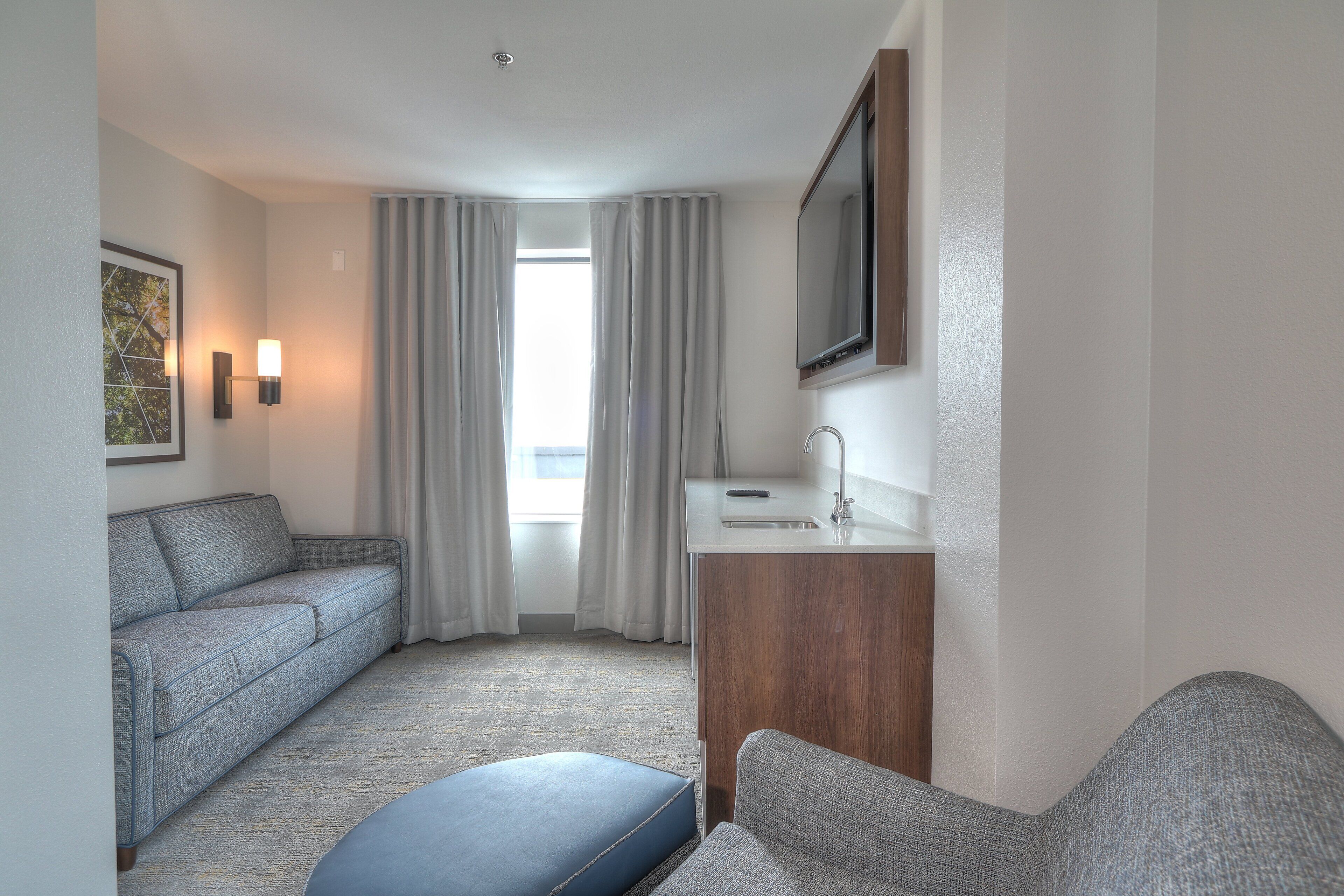 A hotel room with a gray sofa, a blue ottoman, a wooden side table with a mirror, and a window with curtains.