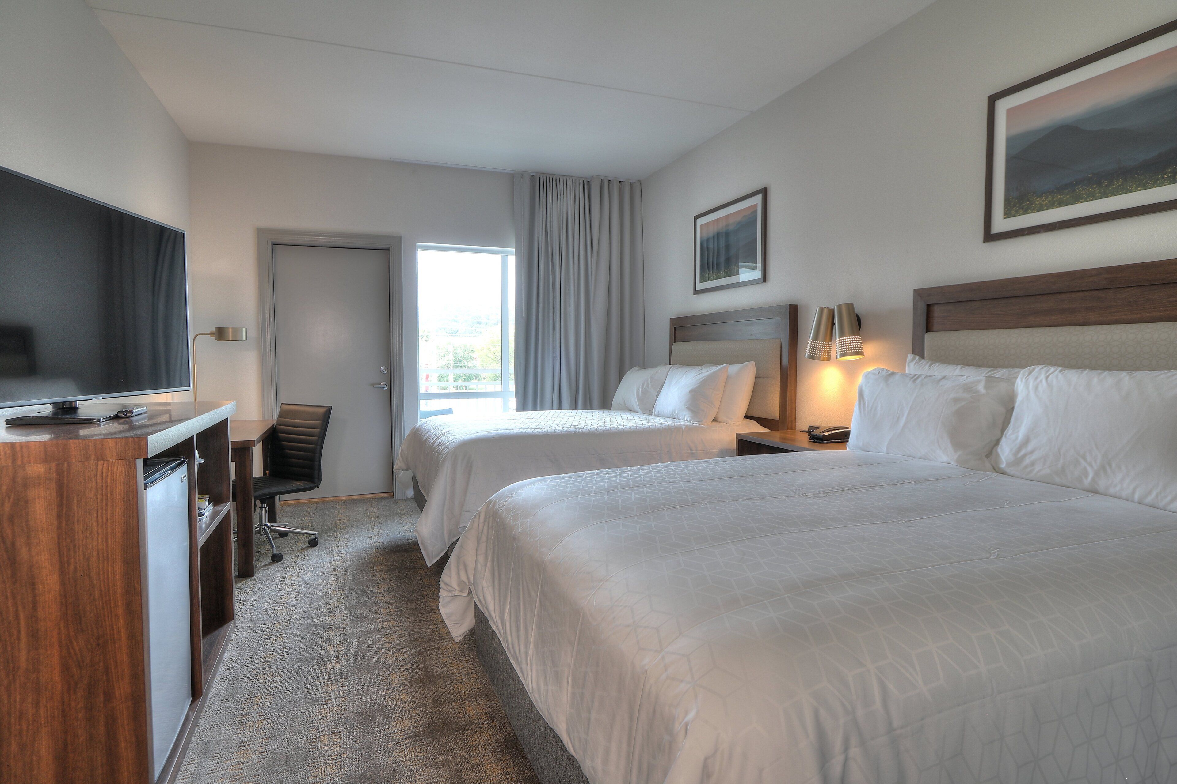 A hotel room with two beds, a desk with a TV, a chair, and a door leading to a balcony.