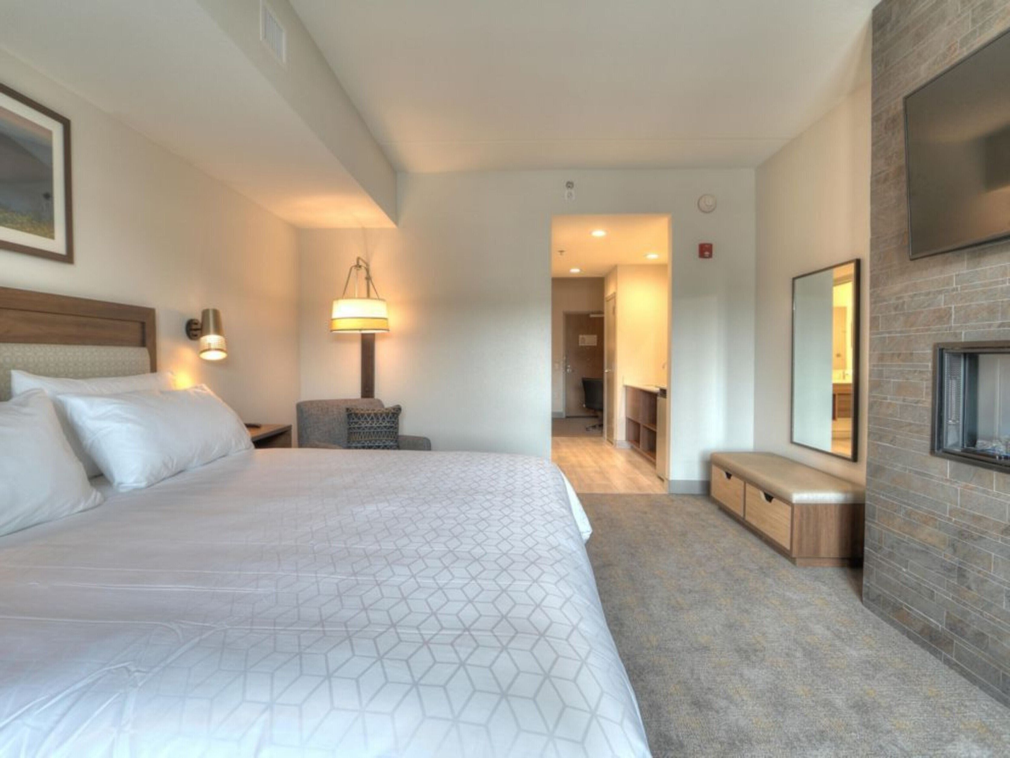 A hotel room with a large bed, a desk, a chair, a lamp, a mirror, and a fireplace.