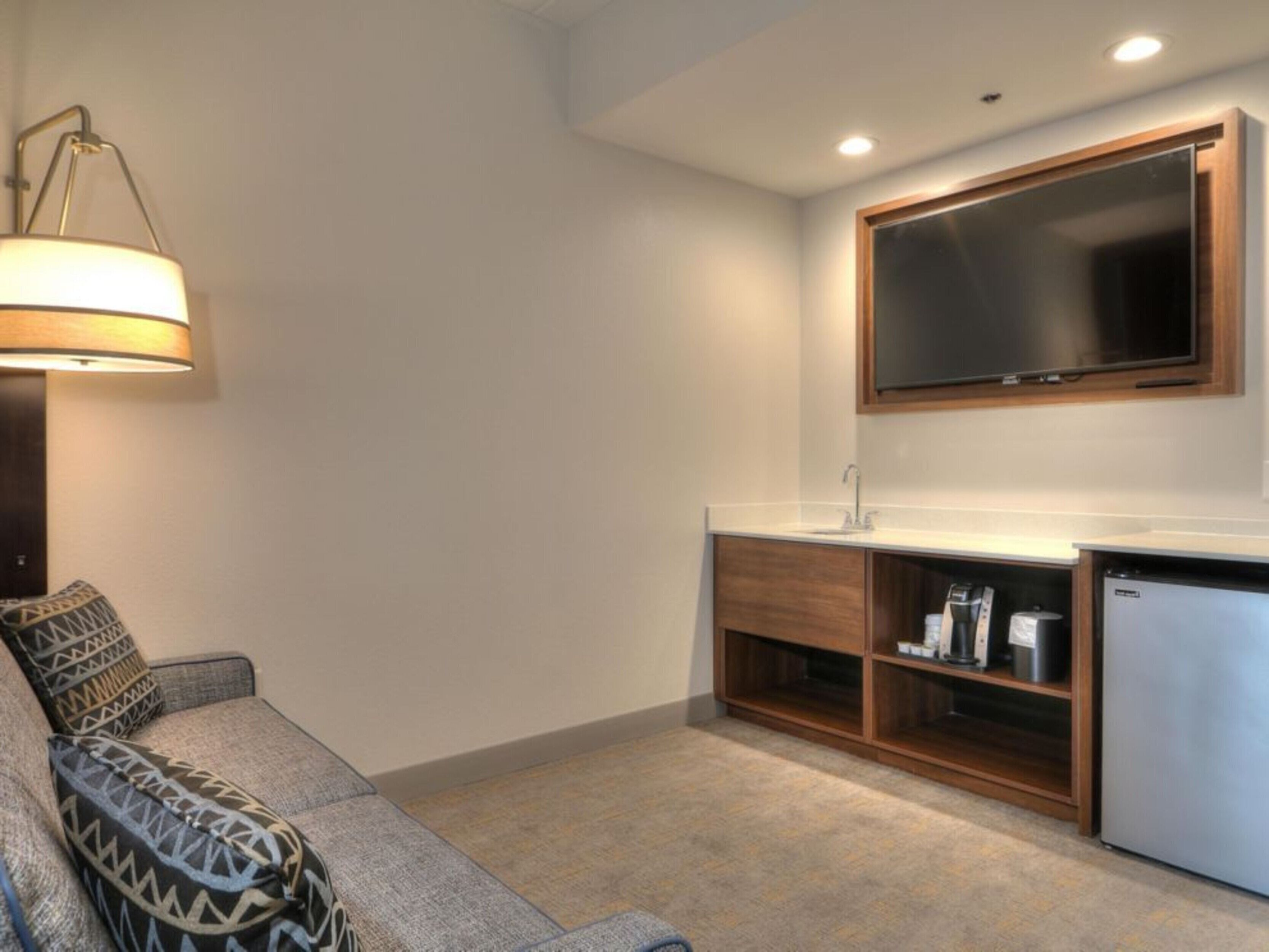 Una sala de estar moderna con un sofá, una mesa de centro, una lámpara, un televisor y una cocina integrada con un lavabo y estanterías.
