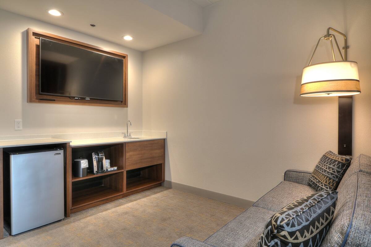 A compact living space with a kitchenette, a flat-screen TV, a sofa with patterned cushions, and a wall-mounted lamp.