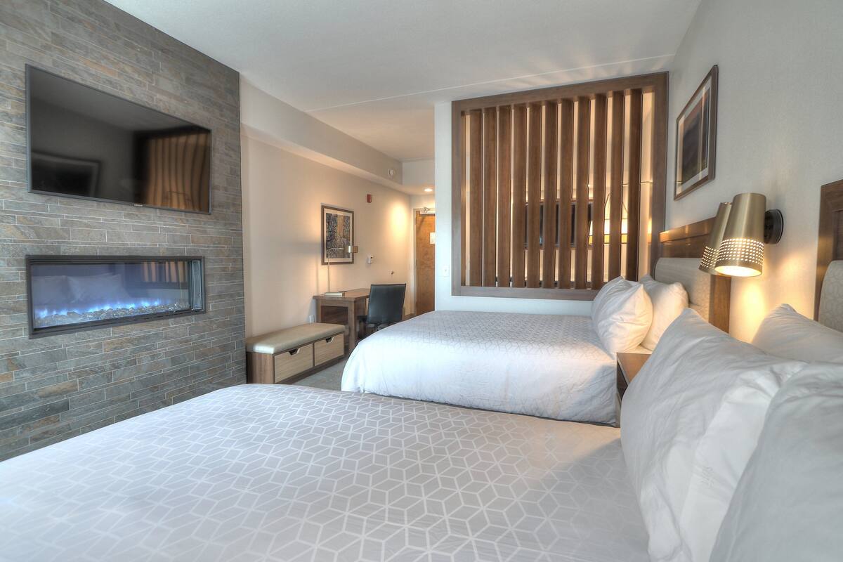 A hotel room with two beds, a fireplace, a desk, and a large window with blinds.