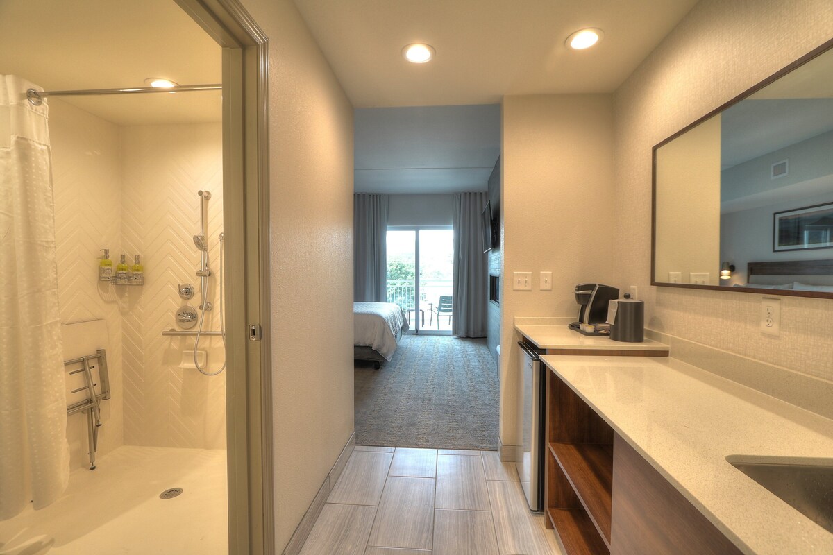 A modern hotel bathroom with a shower, toilet, and sink. There is a bed visible in the adjacent room.
