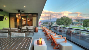 Un área de estar moderna al aire libre con mesas y sillas, un elemento decorativo de fuego y vistas a un estacionamiento y un paisaje distante.
