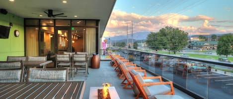 Un área de estar moderna al aire libre con mesas y sillas, un elemento decorativo de fuego y vistas a un estacionamiento y un paisaje distante.