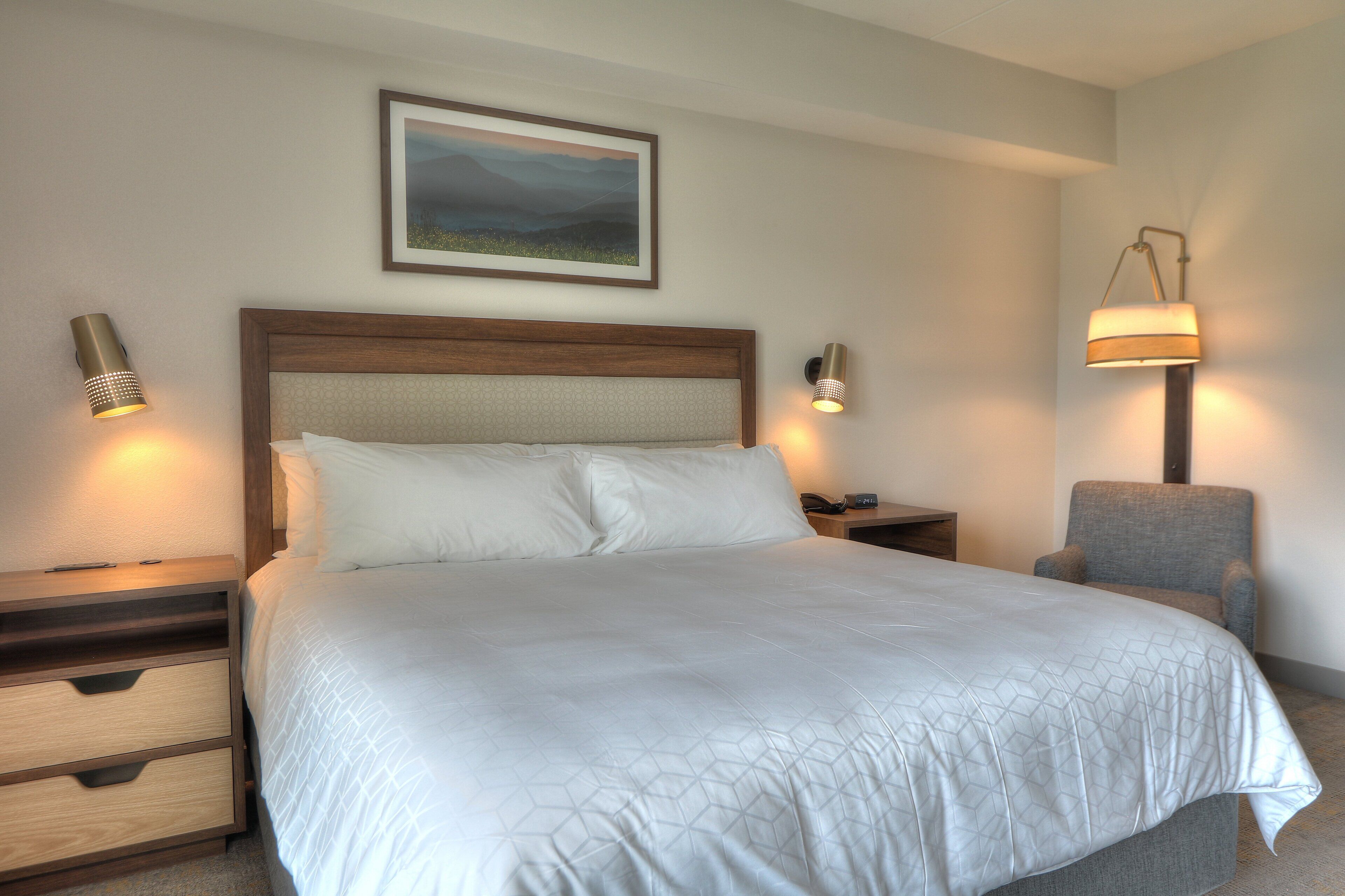 Una cama con sábanas bien puestas, cabecera de madera, dos mesitas de noche con lámparas, un sillón gris y un cuadro en la pared.