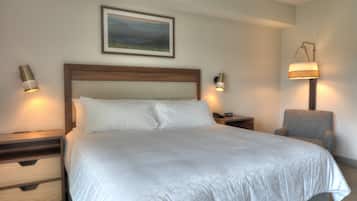 Una cama con sábanas bien puestas, cabecera de madera, dos mesitas de noche con lámparas, un sillón gris y un cuadro en la pared.