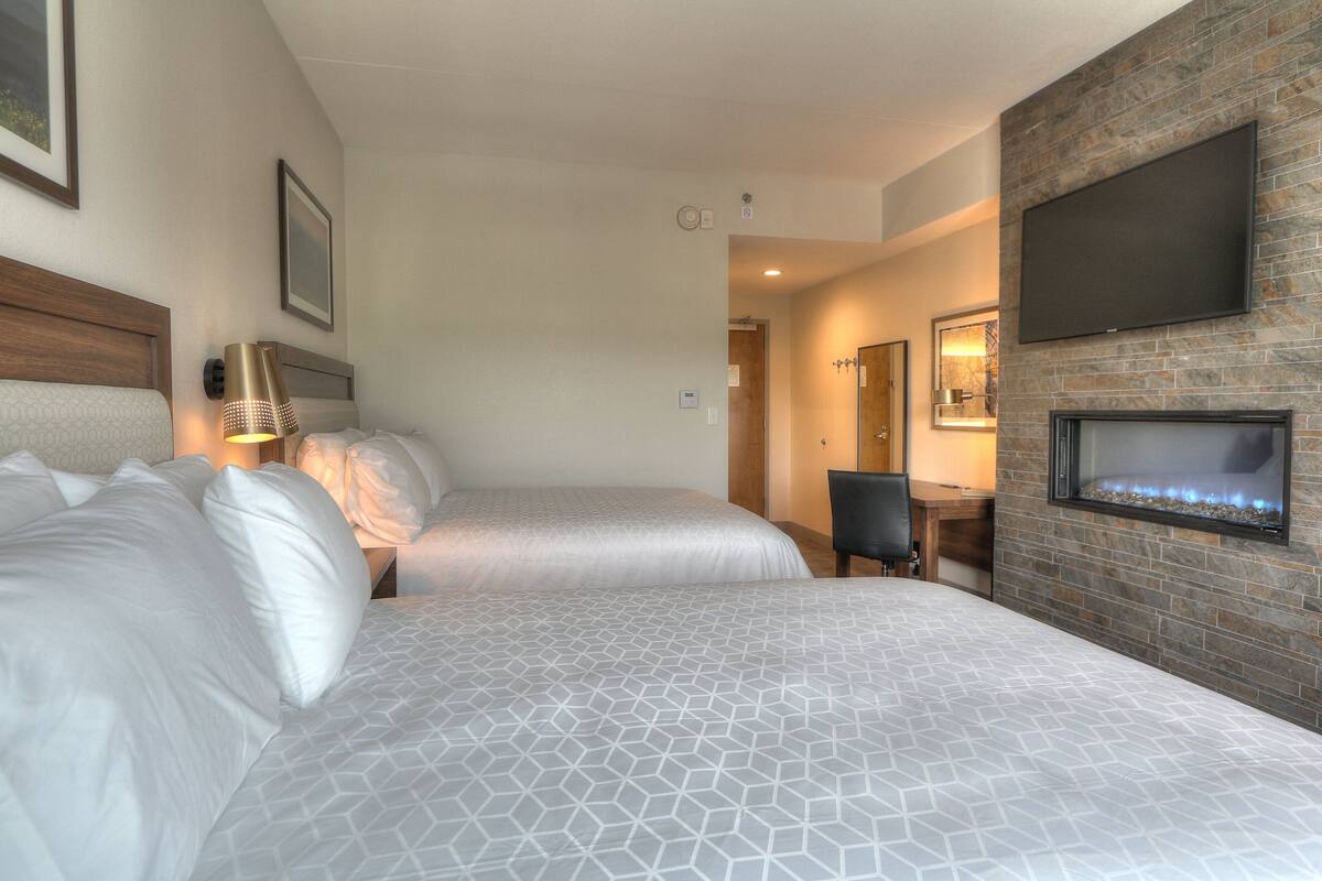 A hotel room with two beds, a fireplace, a TV mounted on the wall, and a desk with a chair.