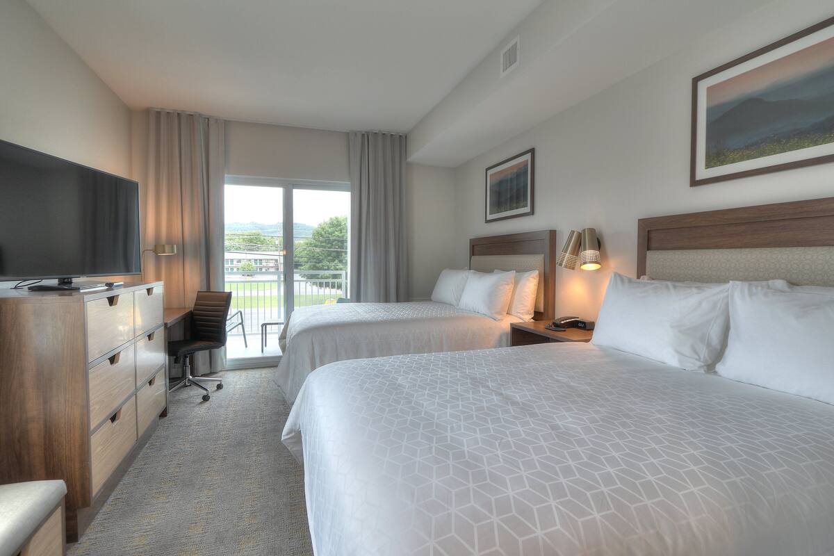 A hotel room with two beds, a desk with a flat-screen TV, a chair, and a large window with curtains.