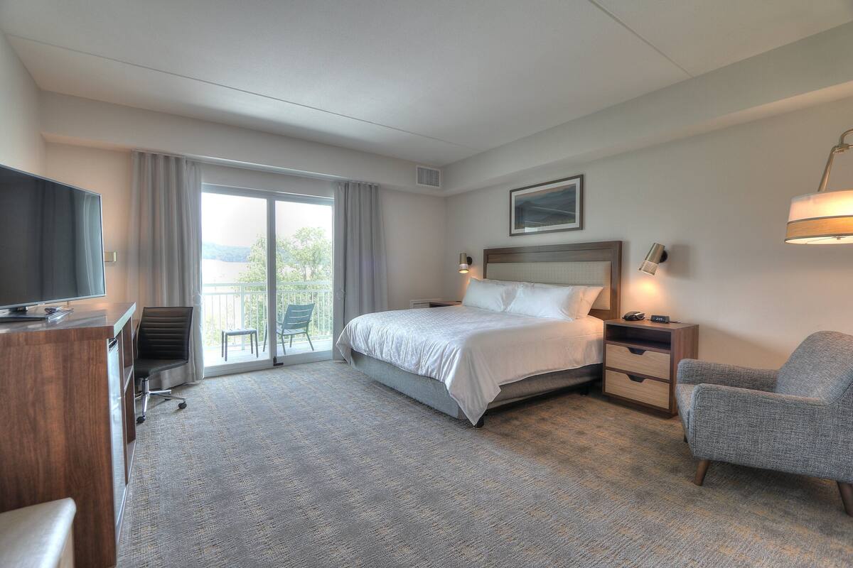 A hotel room with a bed, a desk, a chair, a TV, a lamp, and a view of the outdoors.