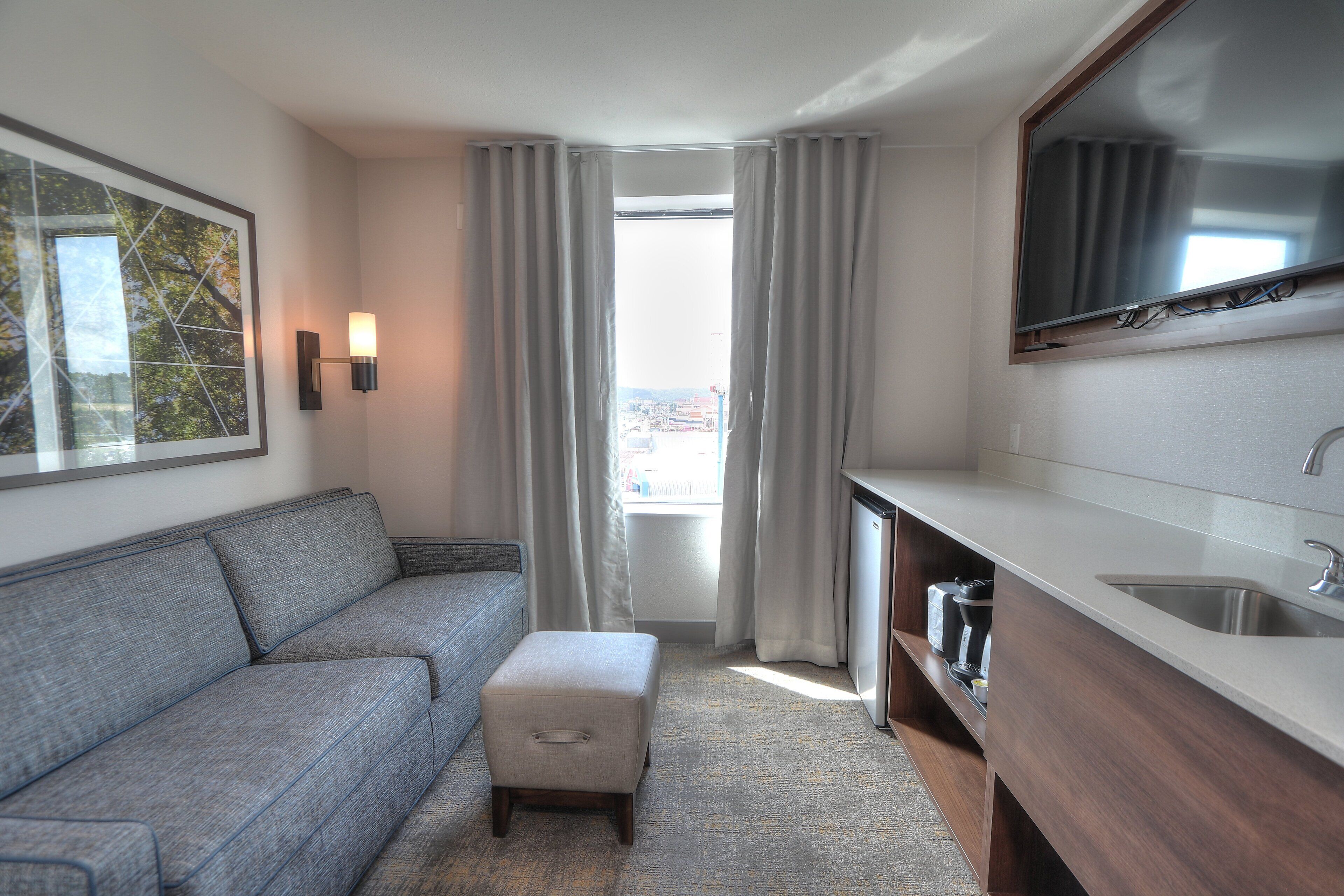 Une chambre d’hôtel moderne avec un canapé gris, un petit pouf, un coin cuisine avec un évier et une grande fenêtre avec des rideaux.
