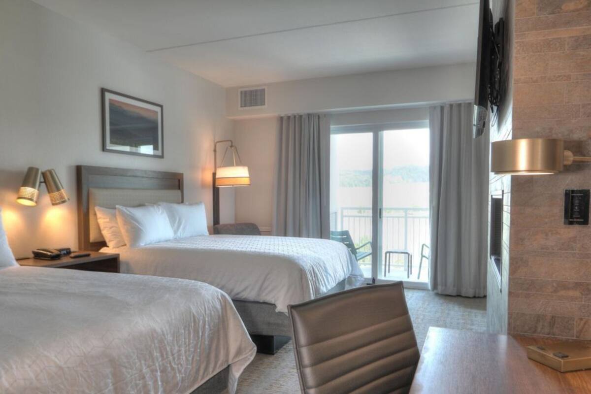 A hotel room with two beds, a desk, a chair, a TV, and a balcony with a view.